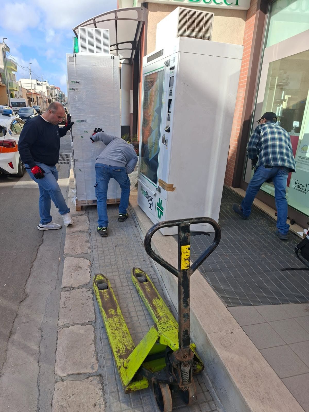 Due operai scaricano elettrodomestici bianchi e alti da un transpallet su un marciapiede cittadino.