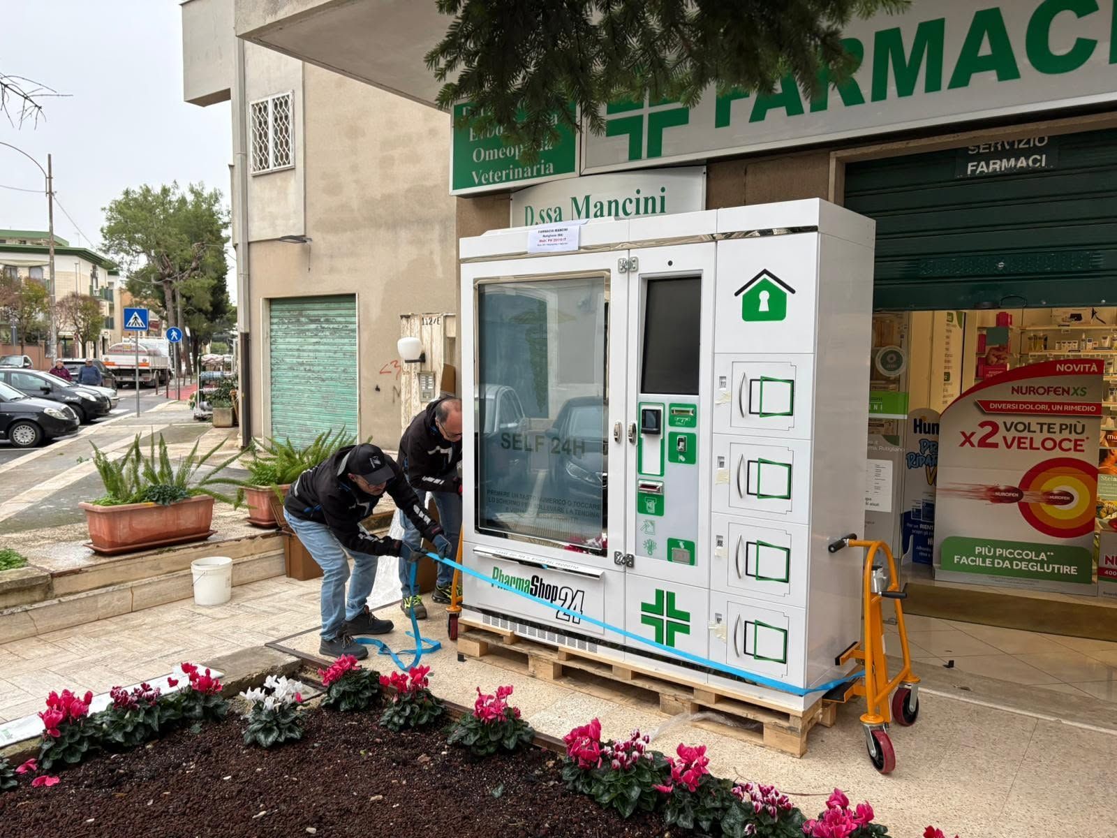 Alcuni operai installano un frigorifero medico bianco all'esterno di una farmacia.