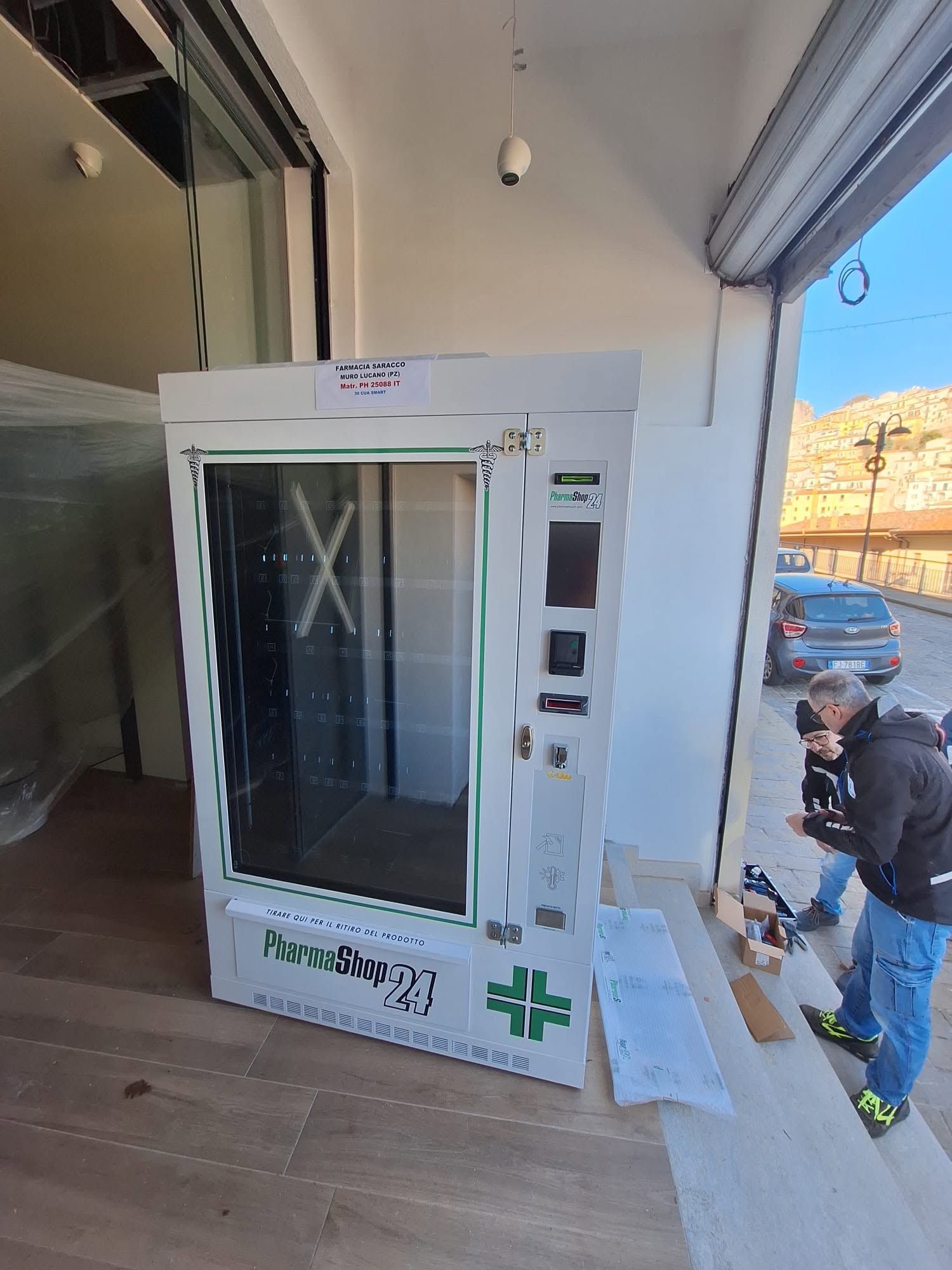 Distributore automatico bianco accanto a una vetrina, con un addetto accovacciato vicino al marciapiede antistante.