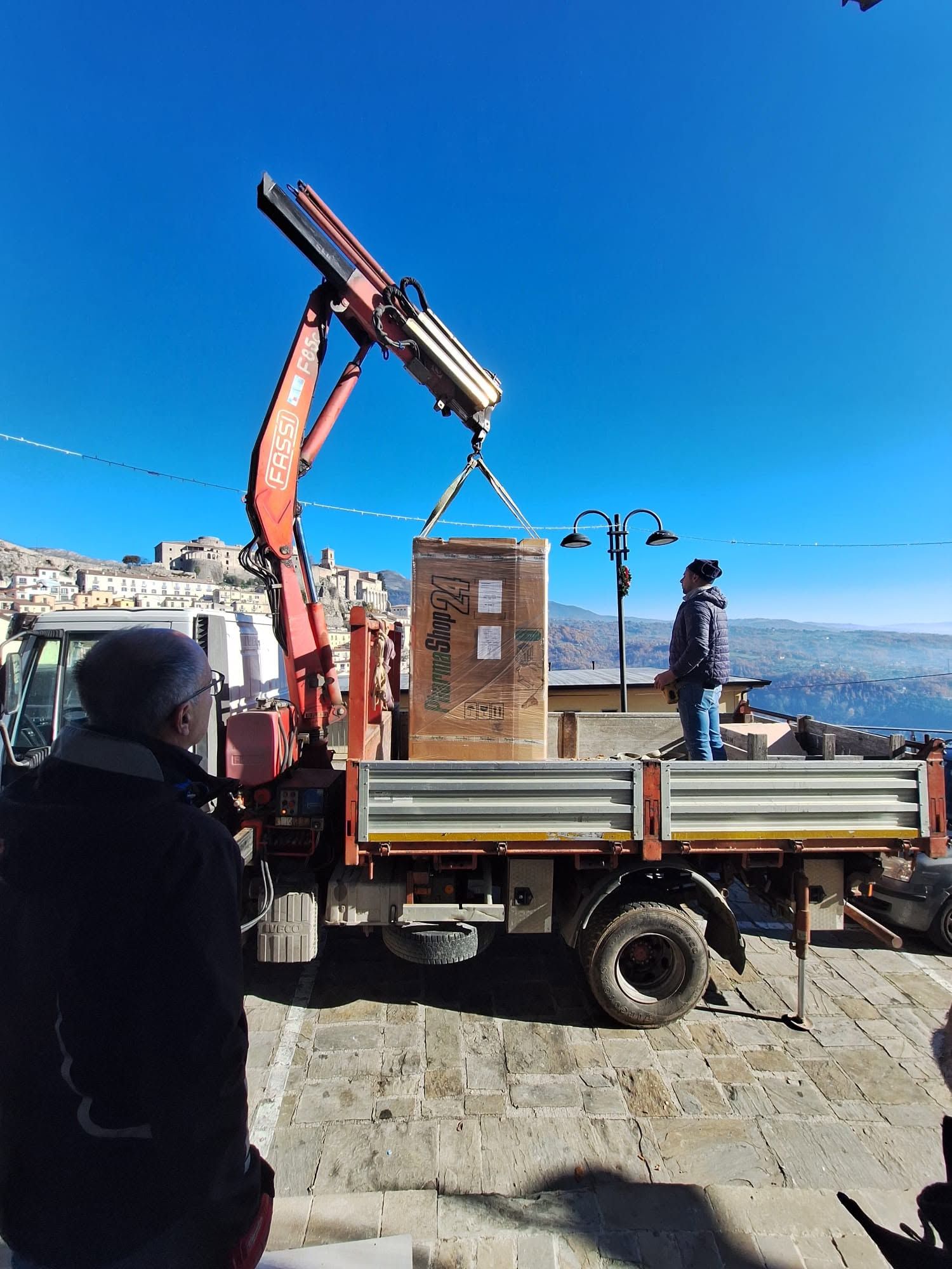 Un camion a pianale con gru scarica una cassa sul lungomare, con operai nelle vicinanze e un cielo azzurro sopra di loro.