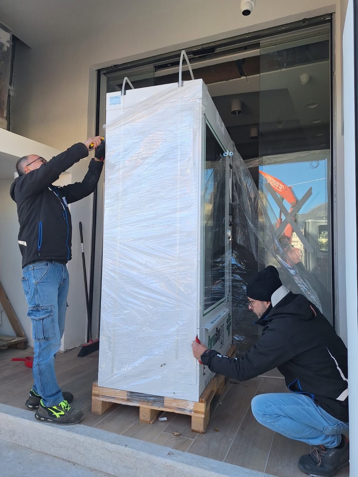 Due operai stanno avvolgendo un grande elettrodomestico bianco in plastica su un pallet di legno vicino a una porta.