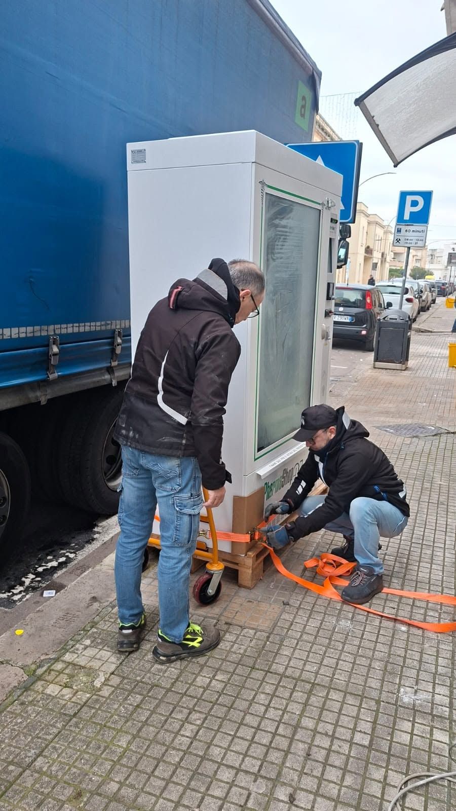 In una zona urbana, alcuni operai fissano con delle cinghie un elettrodomestico bianco su un marciapiede accanto a un camion blu.