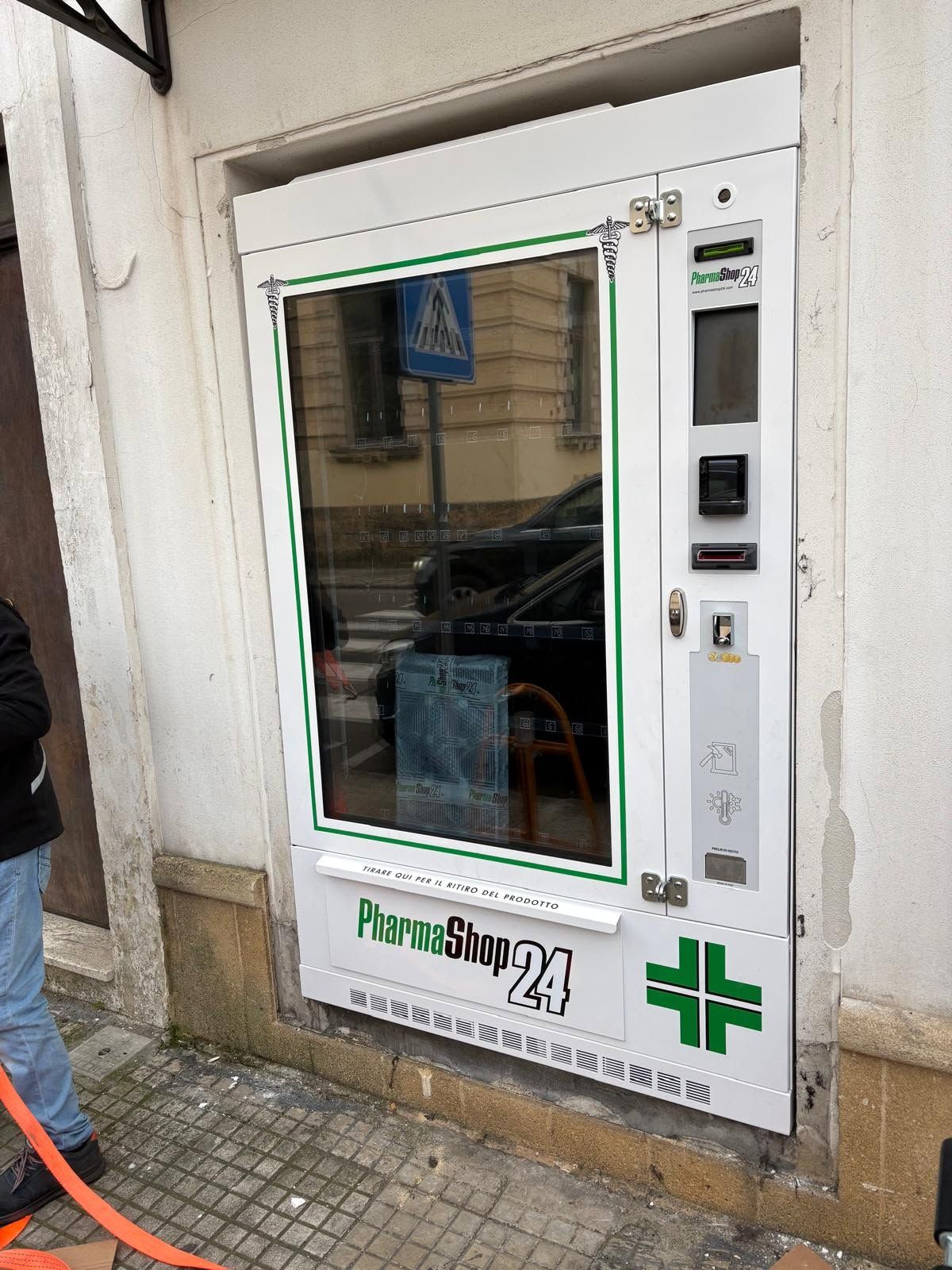 Sportello del servizio farmacia con porta a vetri, fessura per il pagamento e insegna a croce verde sulla facciata dell'edificio.