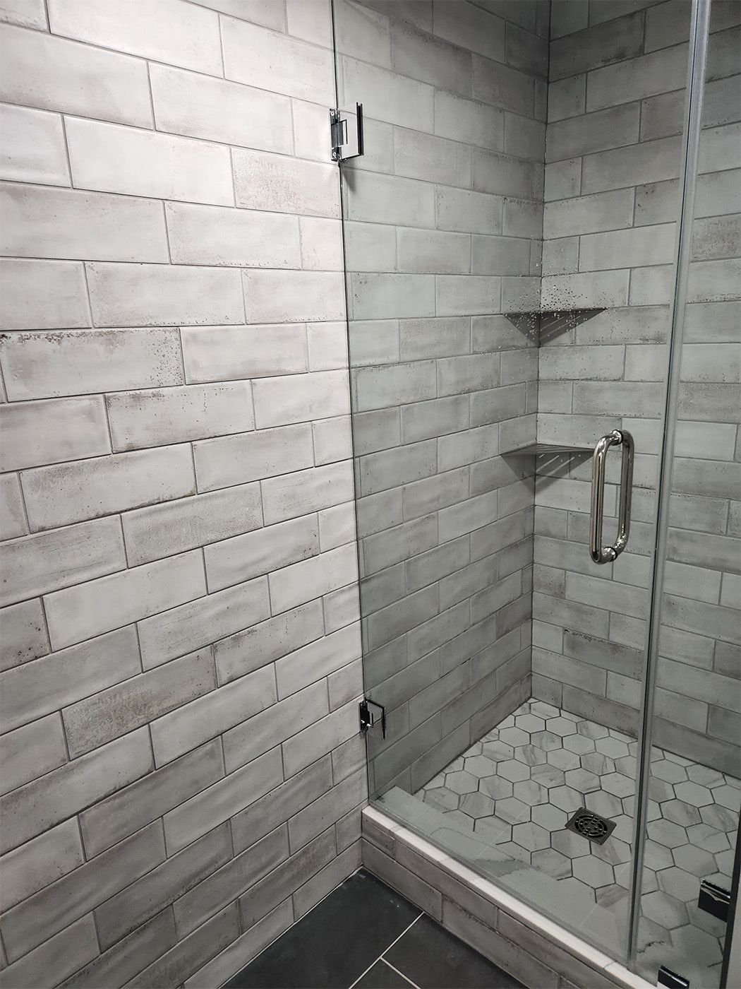 Small tiled shower stall with glass door, built-in corner shelves, and a grab bar.