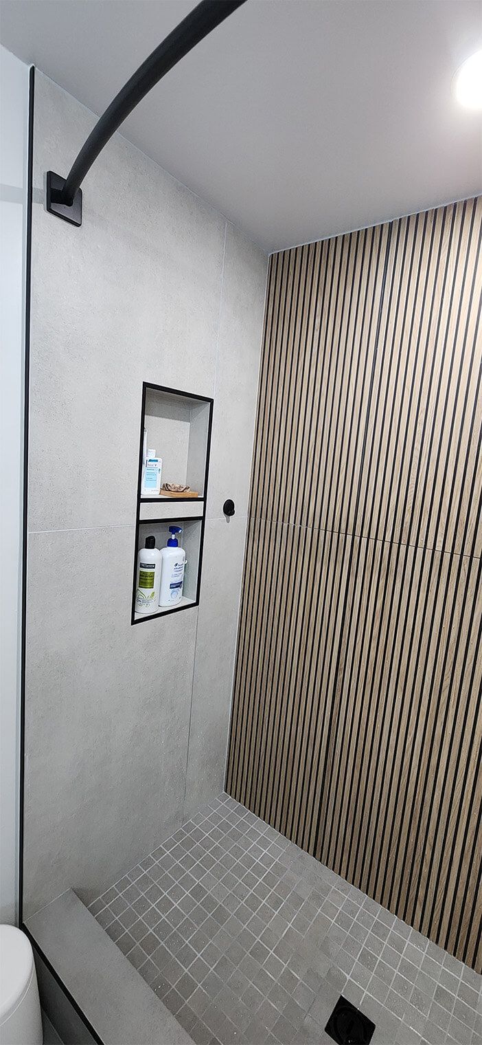 Small tiled shower stall with patterned accent wall, showerhead, mirror, and wall-mounted toiletries.