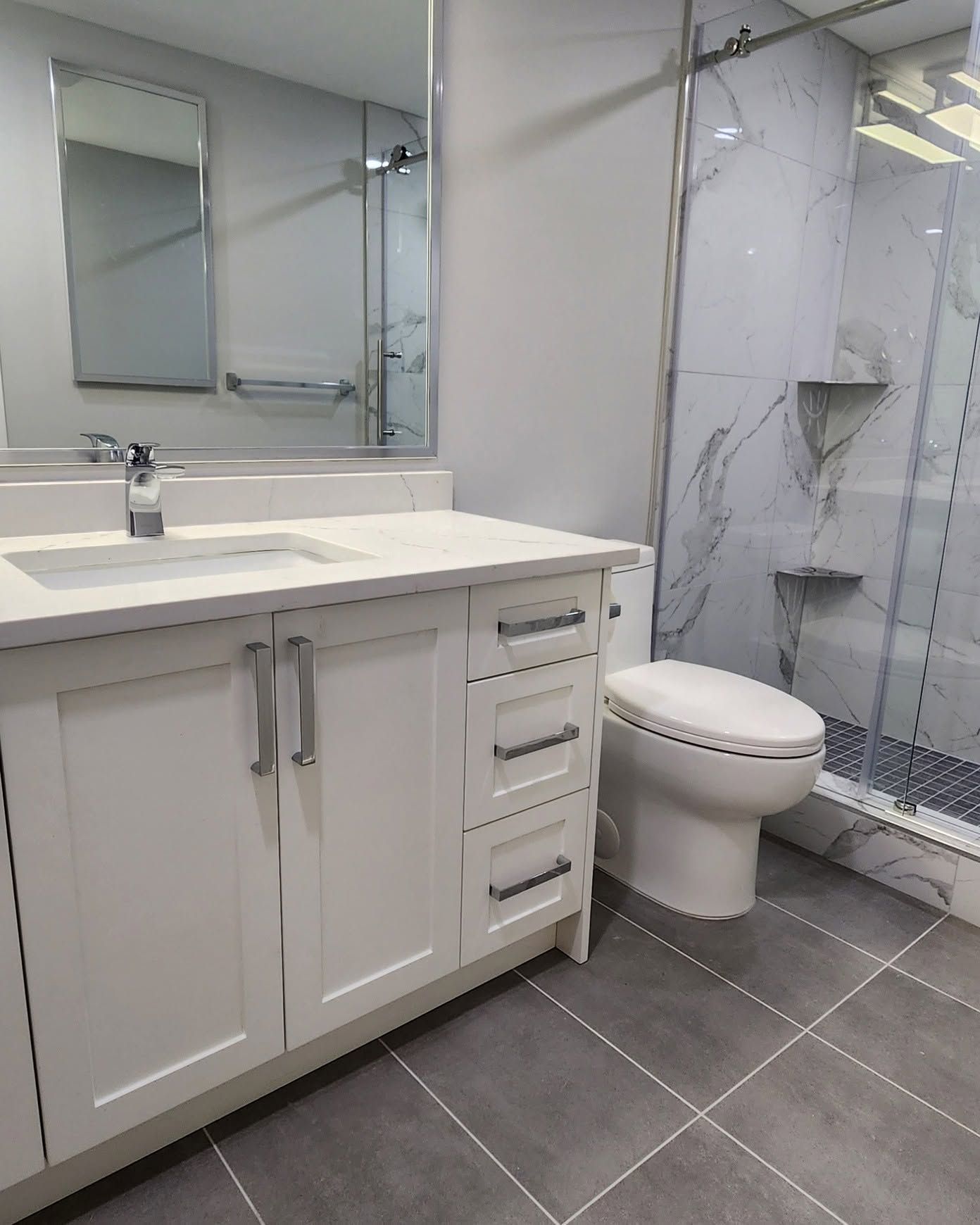 Modern bathroom with white vanity, mirror, toilet, gray tiled floor, and glass shower.