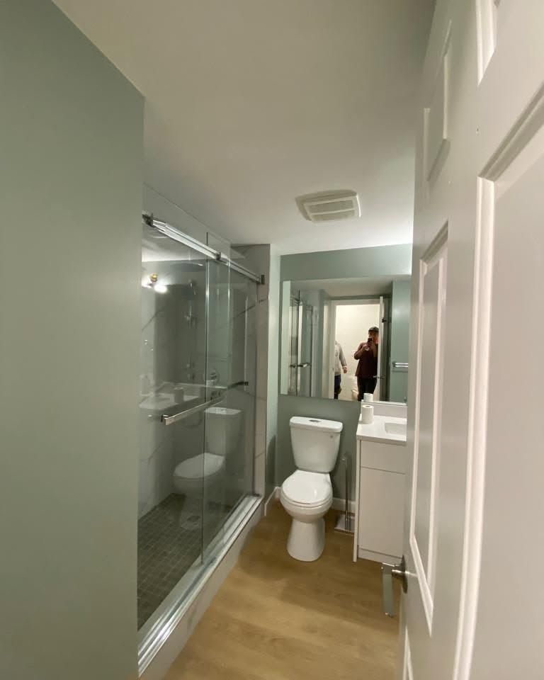 Bright bathroom with glass shower, white toilet, and sink; a person is visible in the mirror.