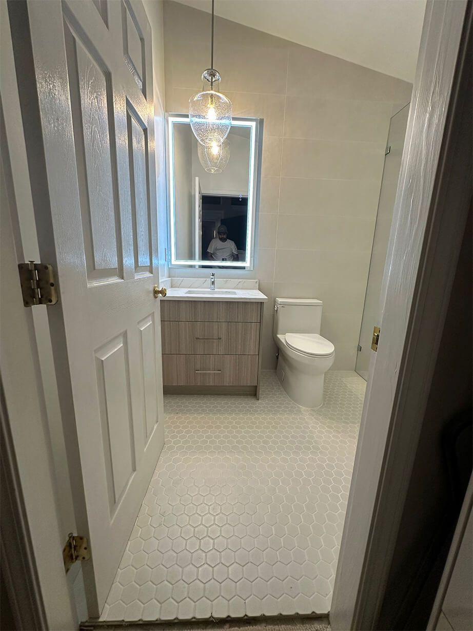 Small bathroom with white vanity, round mirror, toilet, and patterned floor tiles, viewed through an open door