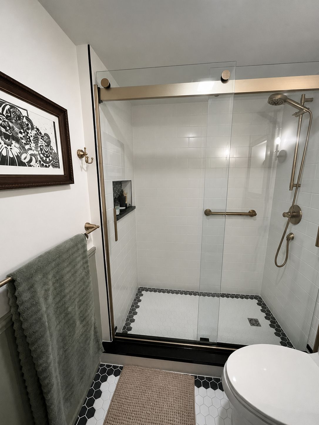 Modern walk-in shower with glass door, black-and-white tile, towel, and white sink in a bright bathroom
