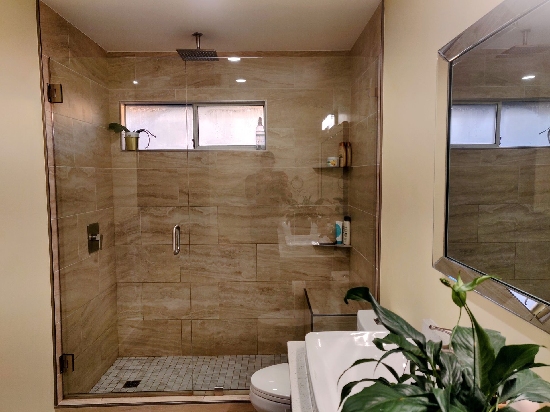 Modern walk-in shower with beige tile, glass enclosure, window, and a potted plant beside a white vanity.