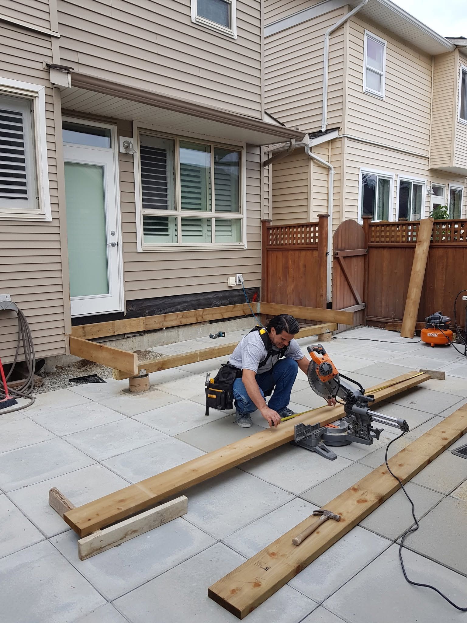 Worker cutting lumber on a patio beside a townhouse, with saws and boards laid out.