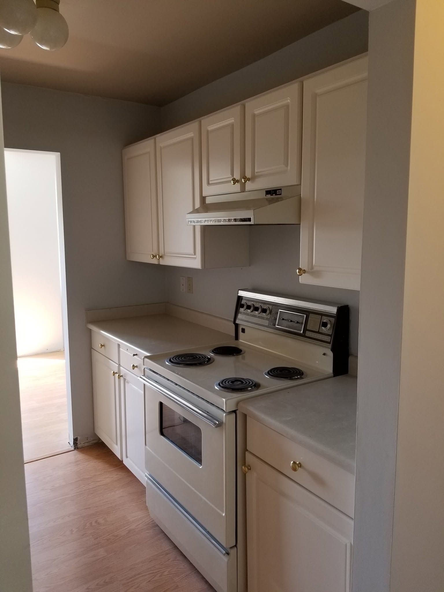 Small white kitchen with cabinets, stove, oven, countertop, and doorway light on the left.