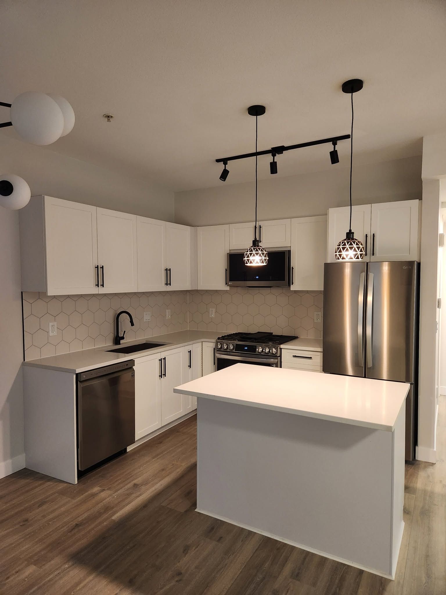 Modern white kitchen with stainless steel appliances, island, and pendant lights on wood-look flooring