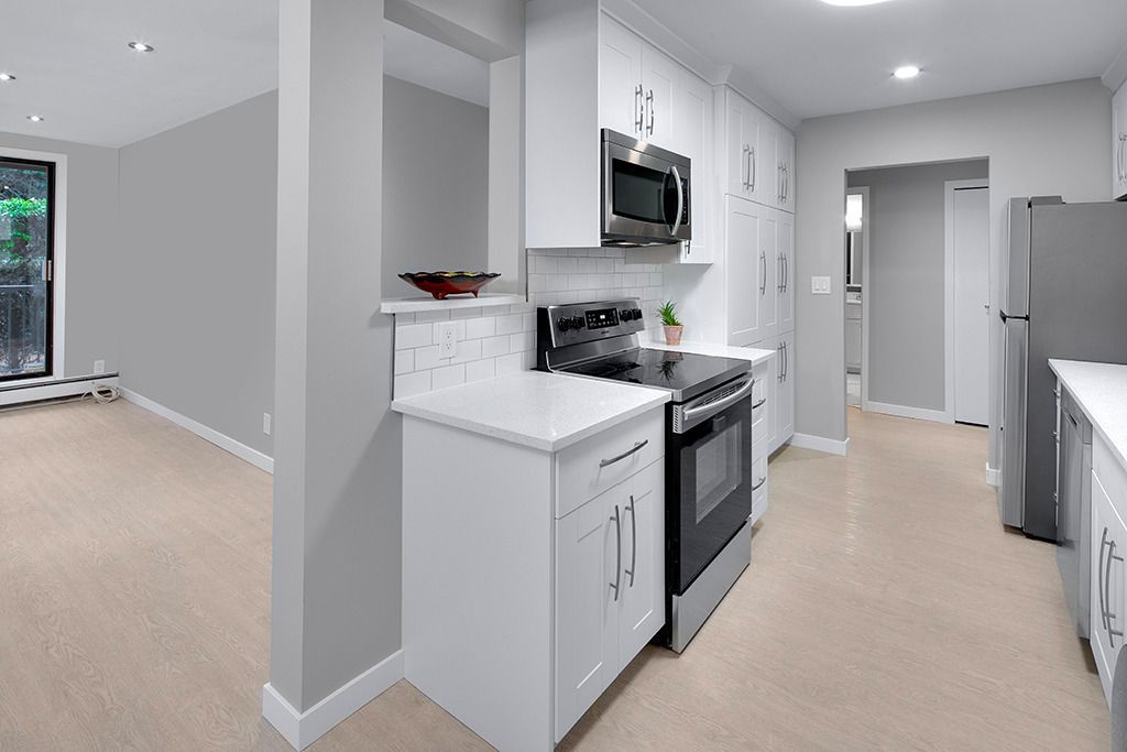Bright kitchen with white cabinets, stainless appliances, and beige carpet opening to a living area