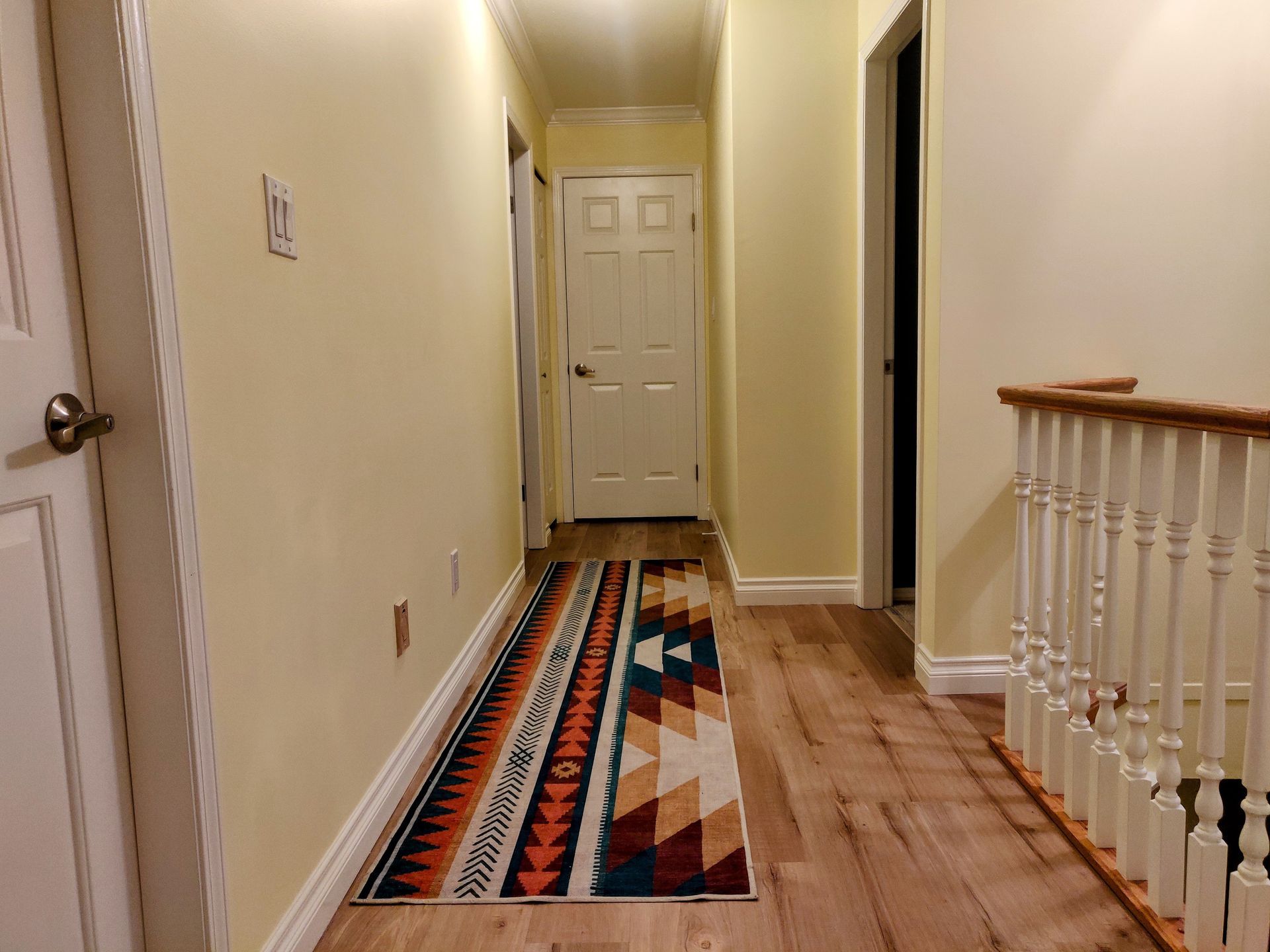 Narrow hallway with a patterned rug, wooden floor, and white door at the end.