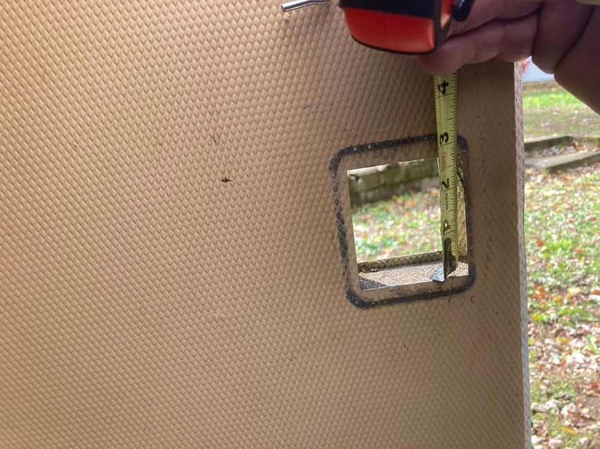 Hand holding a tape measure beside a rectangular cutout in a tan wall panel outdoors