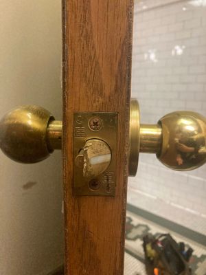 Brass doorknob and keyhole plate on a wooden bathroom door, with a tiled restroom visible behind it.