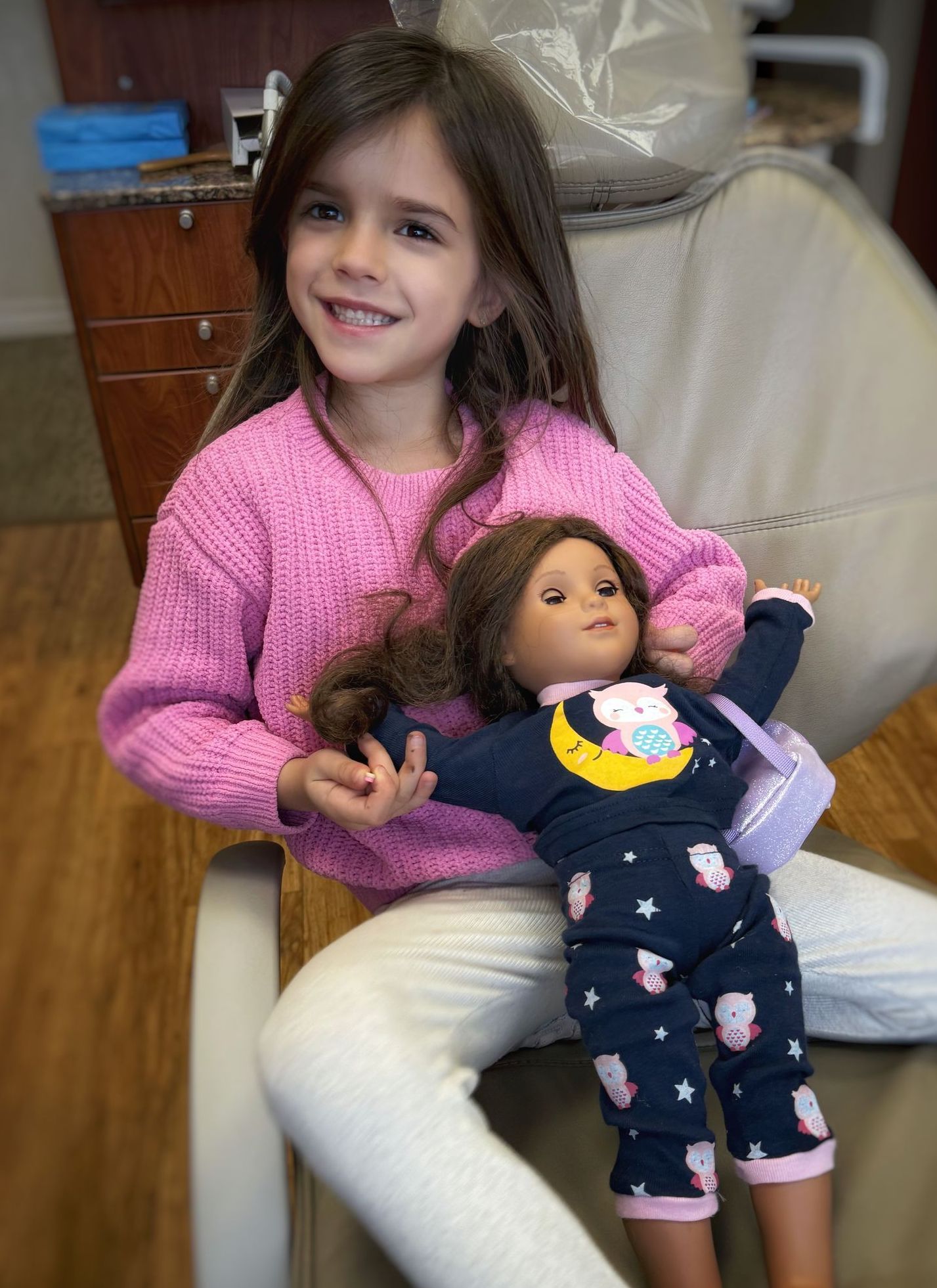 Girl with long brown hair, pink sweater, and doll in pajamas at dentist's office; smiling.