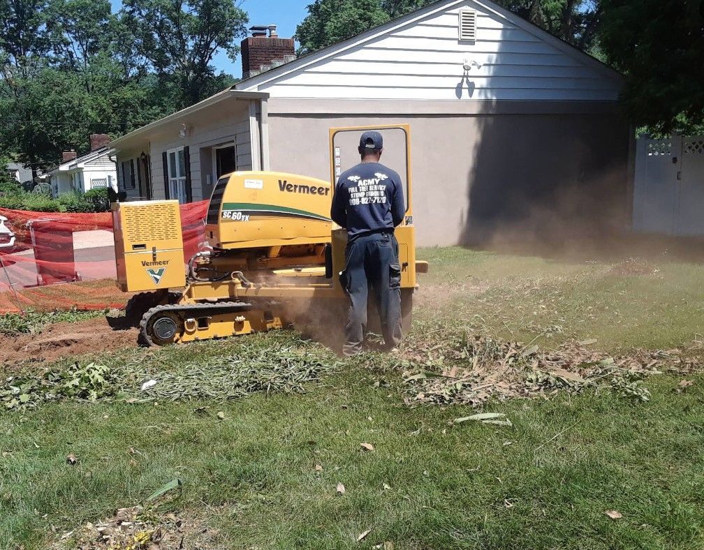 Worker using a yellow Vermeer machine to clear brush beside a house, with dust in the air.