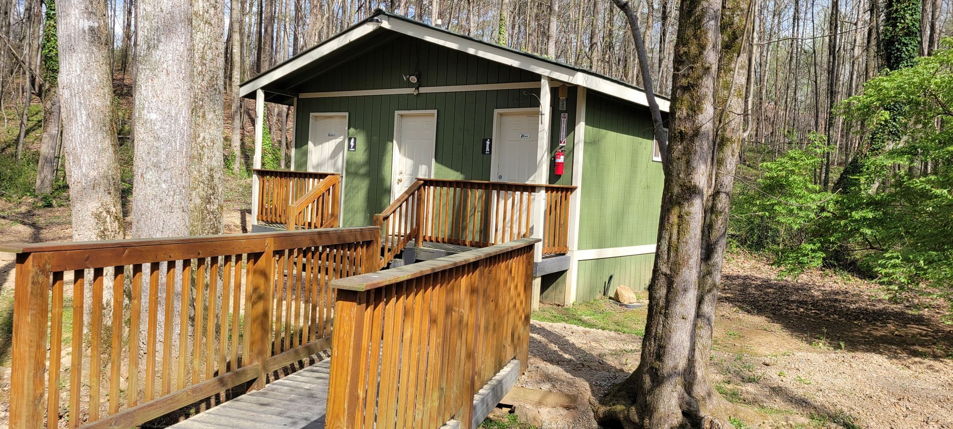 Green building with white doors and a wooden walkway in a wooded area.