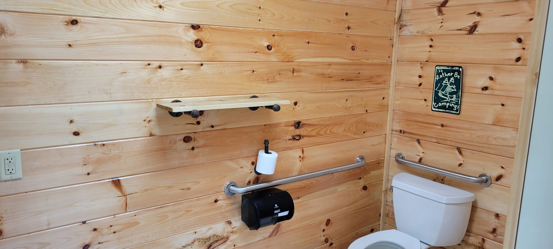 A wooden-paneled bathroom with a toilet, grab bars, and a shelf.