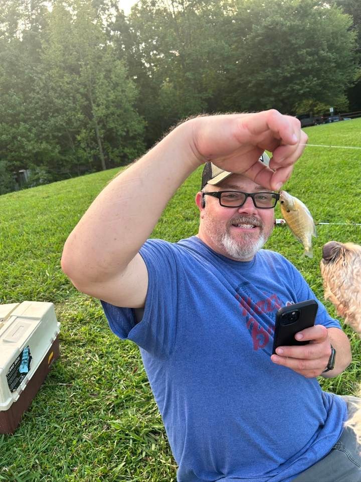 Man holding small fish, smiling while taking a photo on a phone; grassy field and trees in background.
