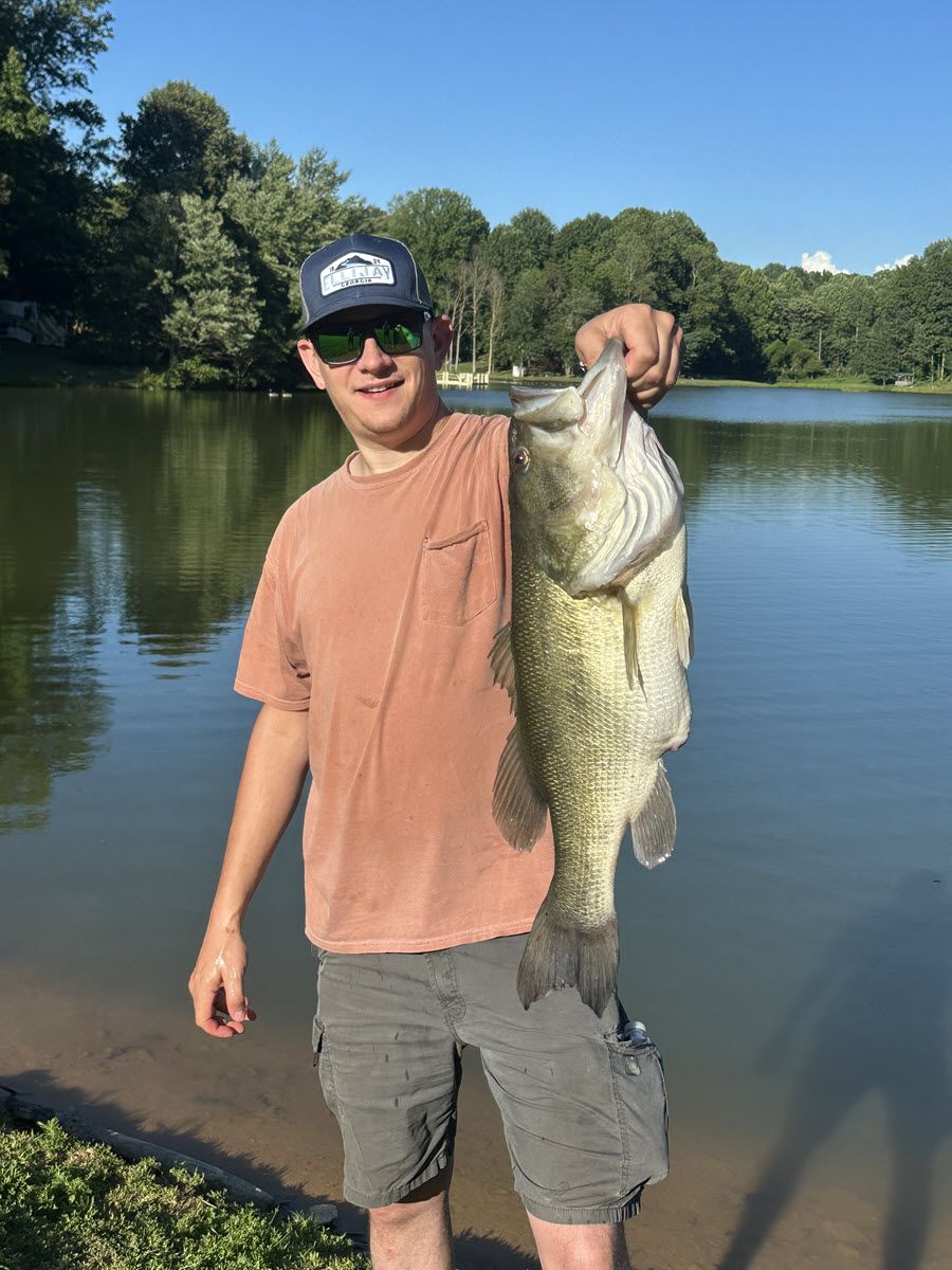 Man holding large bass, standing by a lake. He wears sunglasses, a hat, and shorts.