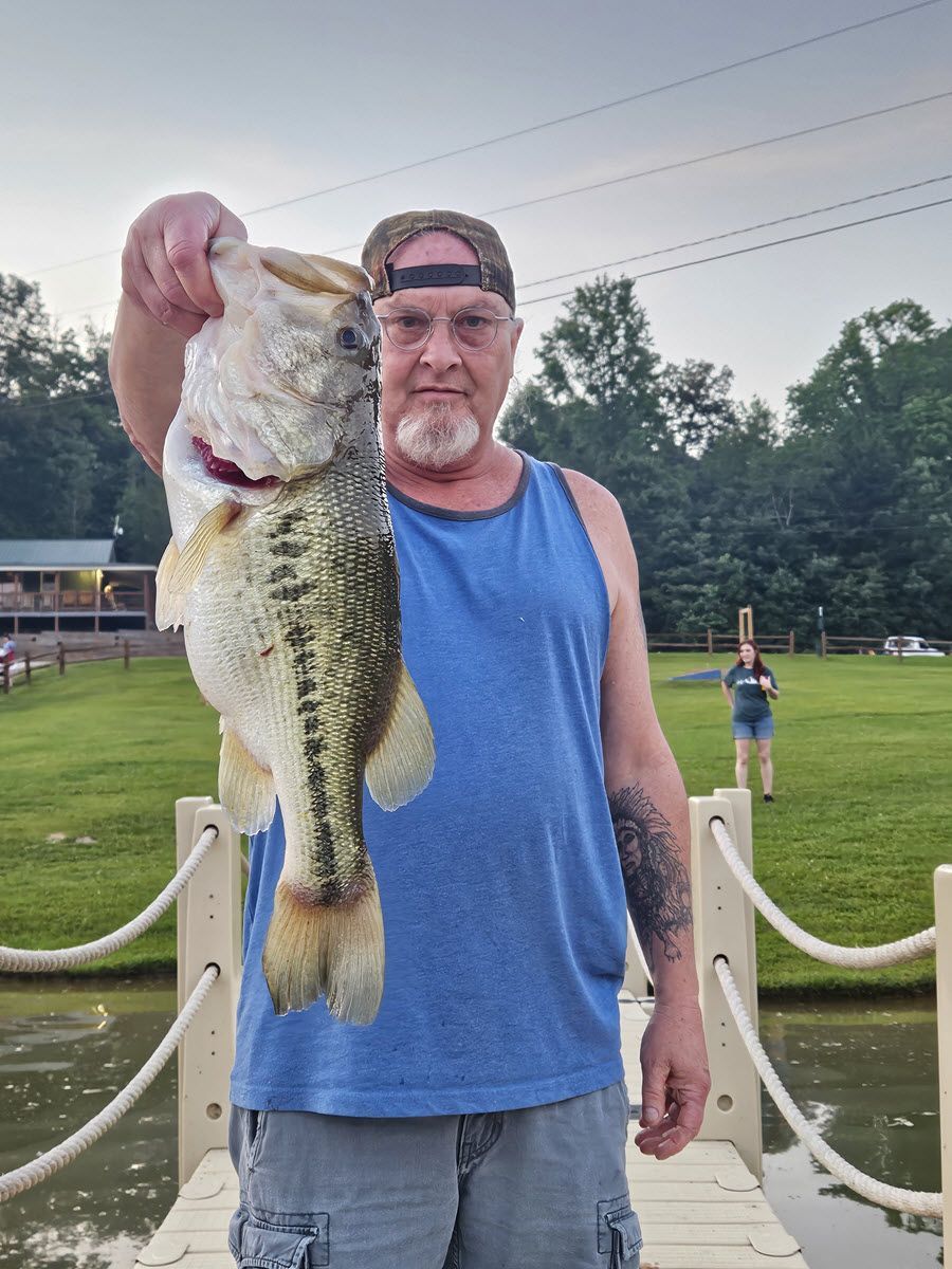 Man holds a large bass fish, posing on a dock. He's wearing a hat and blue tank top, with a woman in the background.