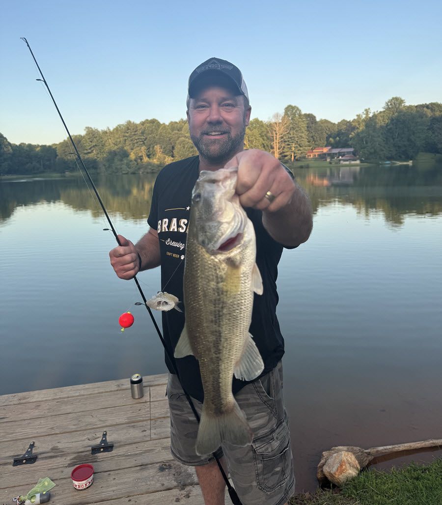 Man holding a large bass he caught while fishing at a lake; wearing a hat, t-shirt and shorts.