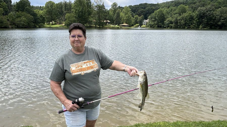Woman holding up a fish she caught, standing by a lake with a fishing rod.