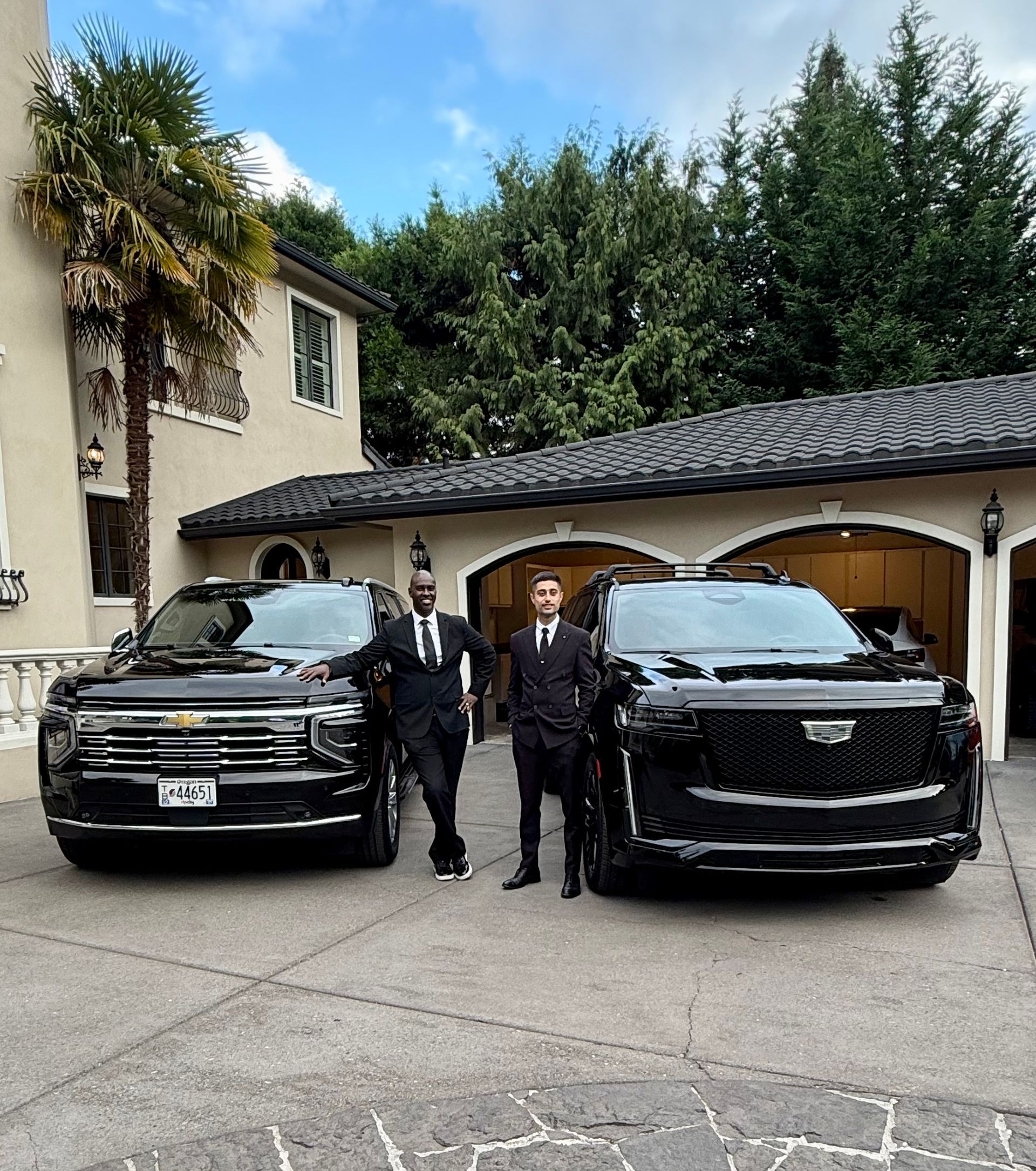 Two chauffeurs in dark suits standing beside a black Chevrolet Suburban and a black Cadillac Escalade in a home driveway