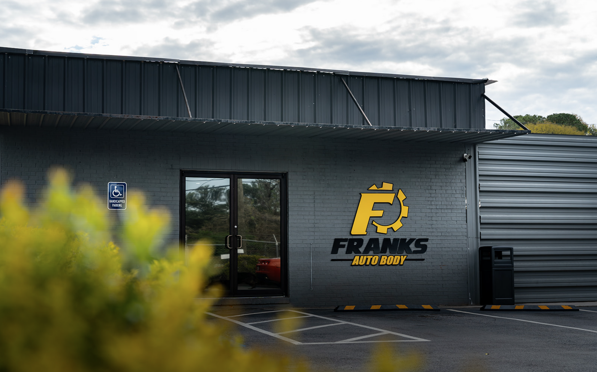 Exterior of a black building with a yellow logo and glass door, framed by shrubs and a gray metal wall.