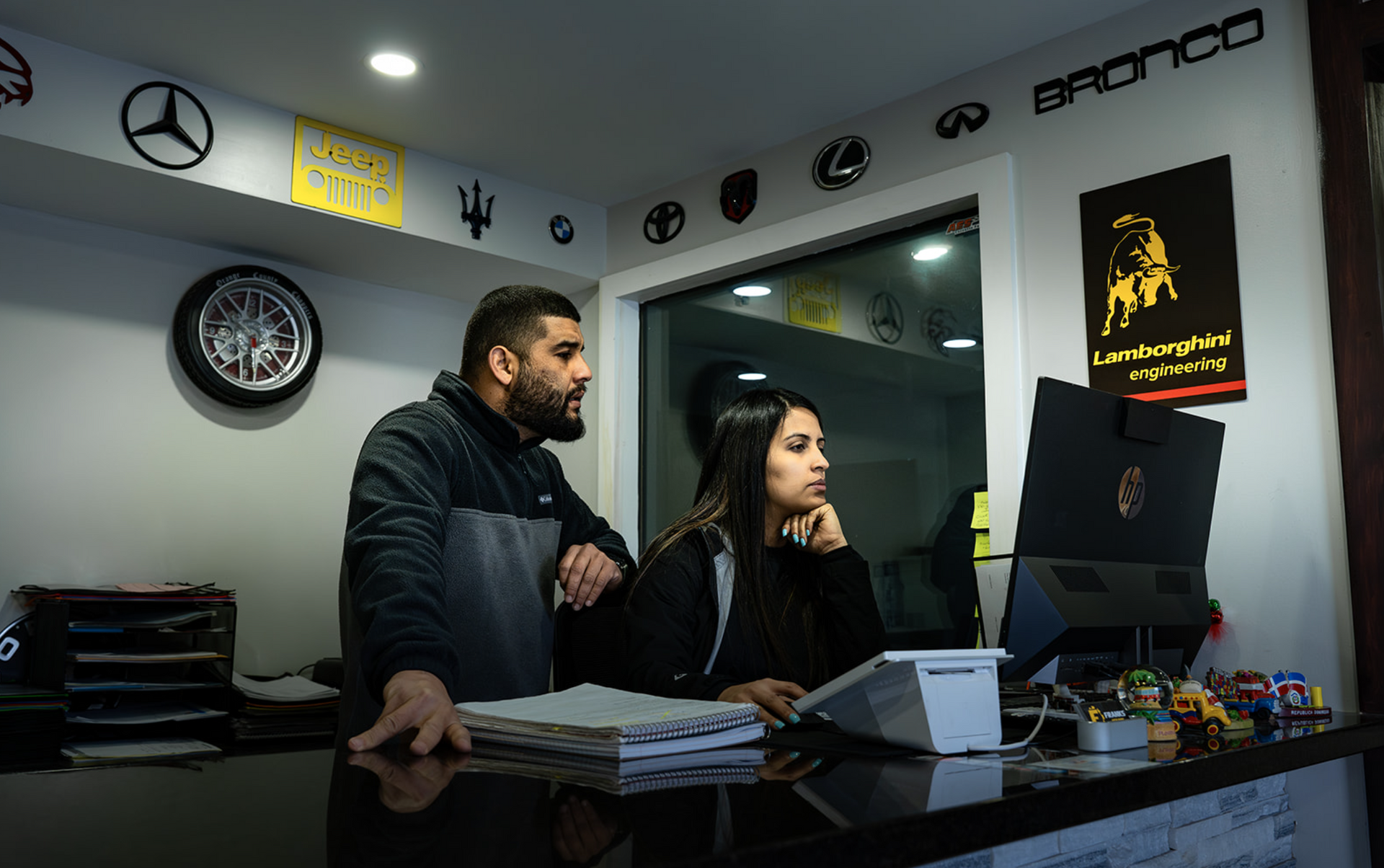 Two people stand at a counter in a modern office, looking at a computer monitor.