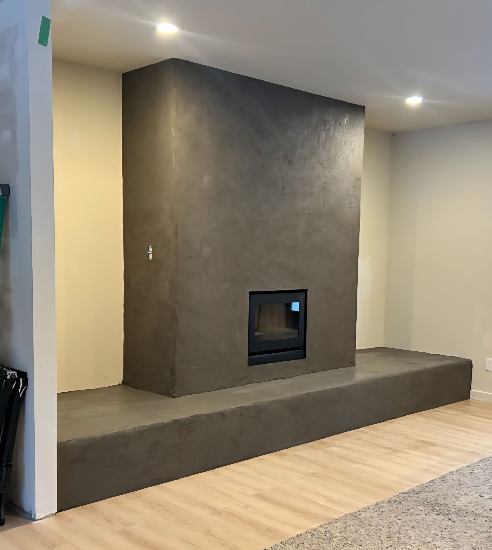 A modern, charcoal-colored concrete fireplace with a central insert, built onto a raised platform in a light-floored room.