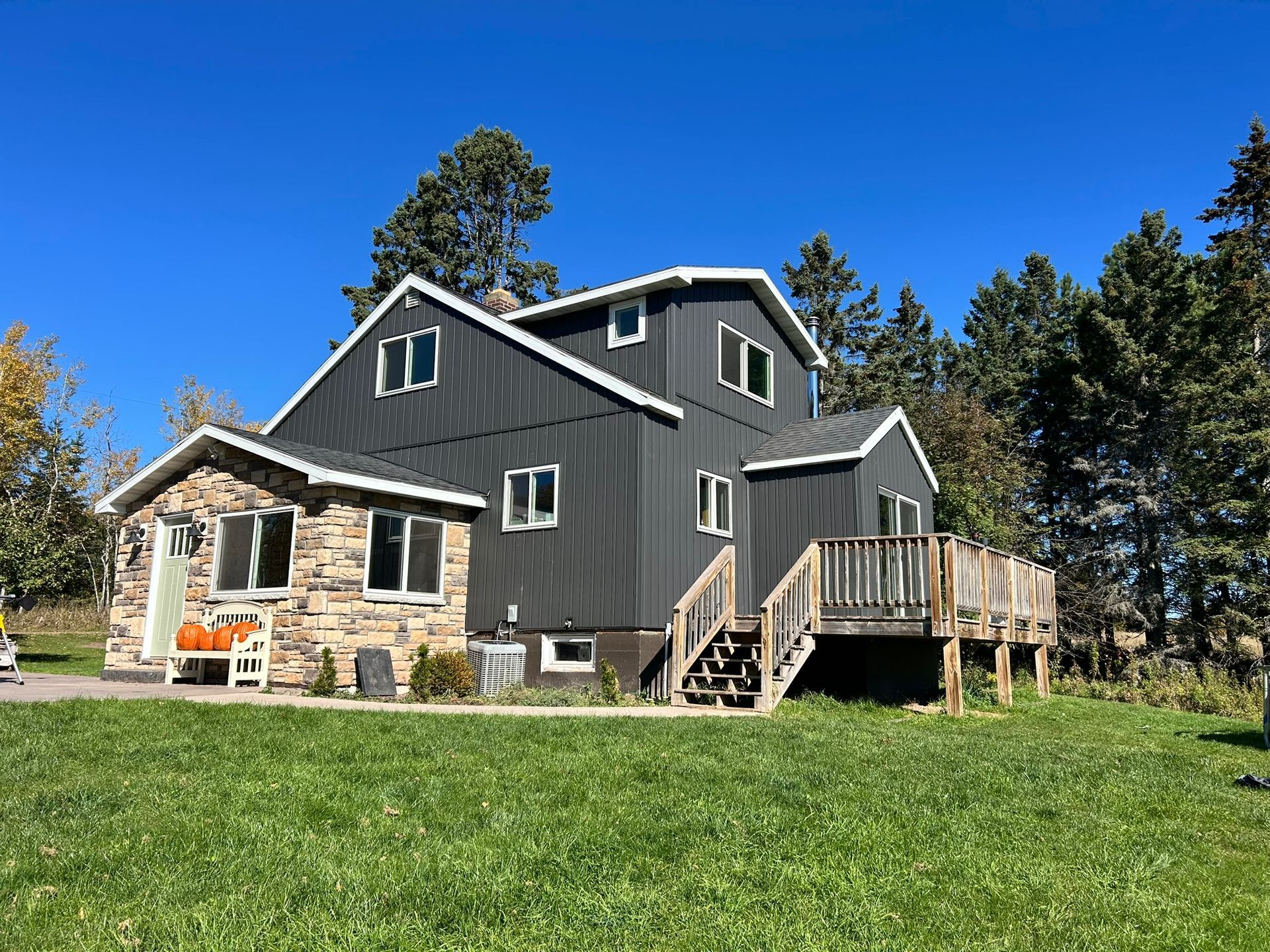 A two-story dark gray home with vertical siding and a stone-accented front entryway, set against trees under a blue sky.