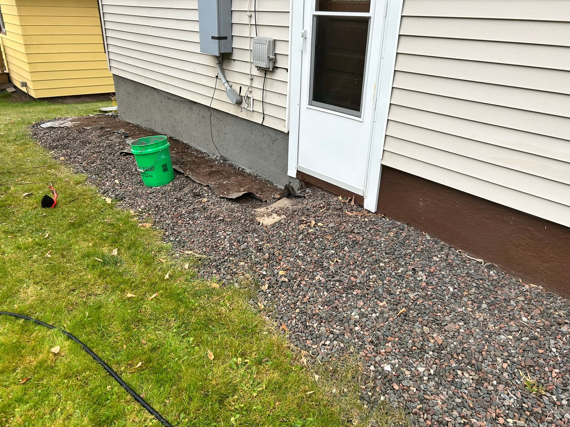 Exterior view of a house wall with beige siding, a white door, and a brown foundation above a gravel landscaping bed.