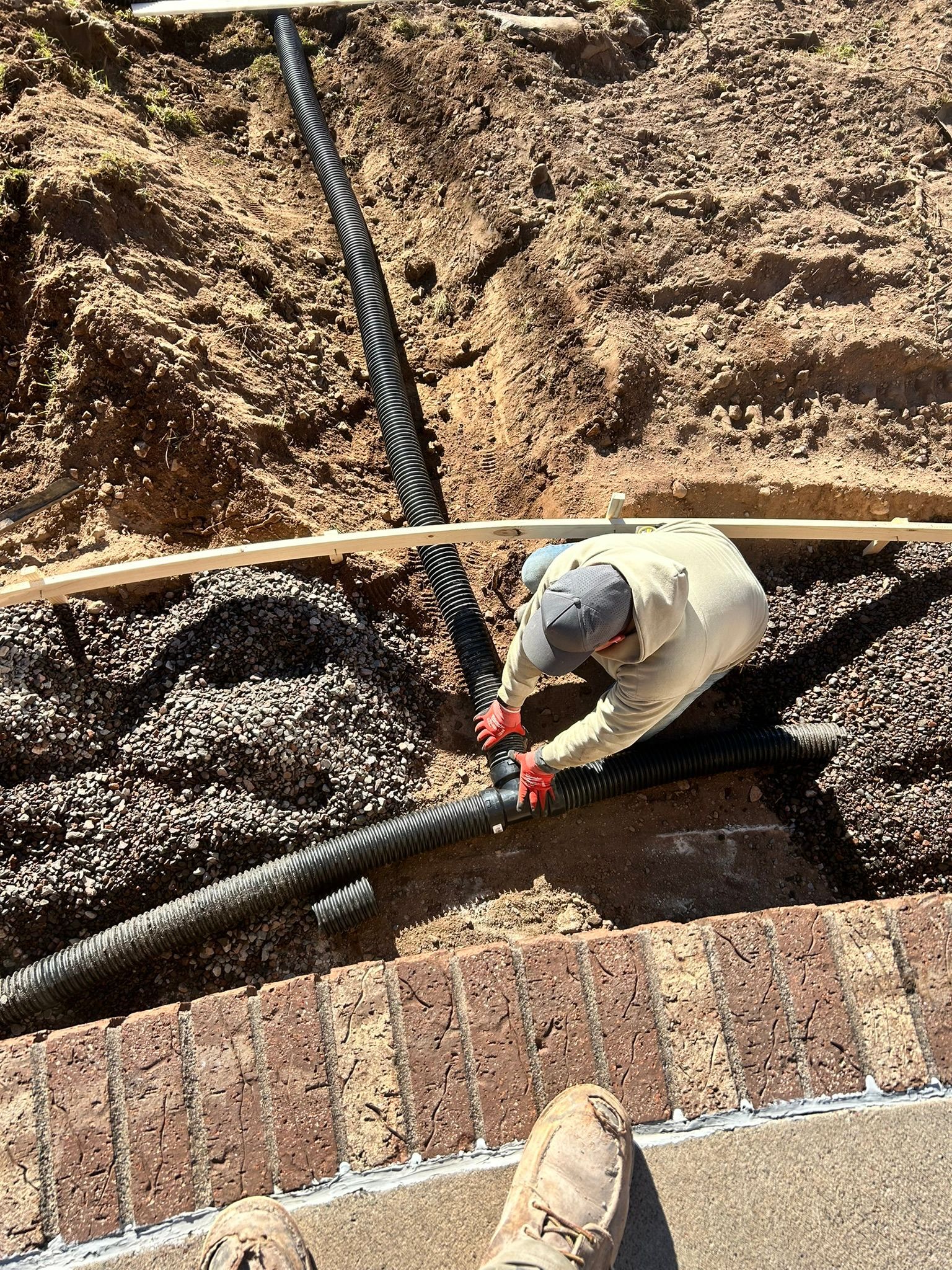 A person in a beige hoodie and red gloves works on connecting black corrugated drainage pipes in a dirt trench.
