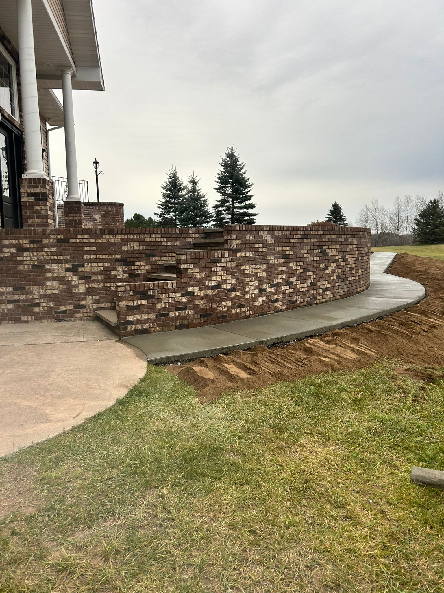 A brick retaining wall and curved concrete walkway bordering a grassy lawn next to a building with white columns.