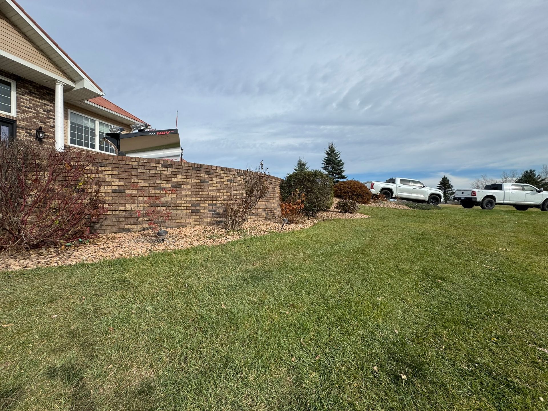 A brick retaining wall sits next to a house with a grass lawn in the foreground and two white pickup trucks in the back.