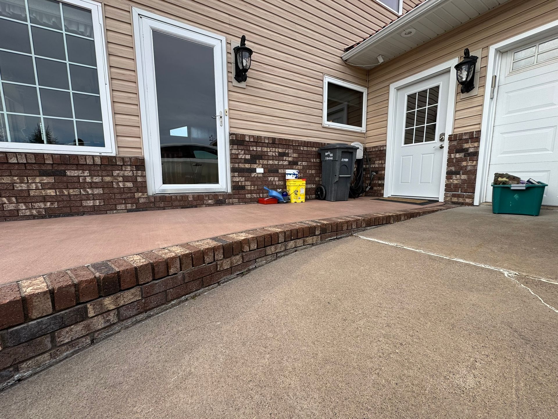 A residential exterior with a brick-trimmed concrete patio, white door and window frames, and beige siding.