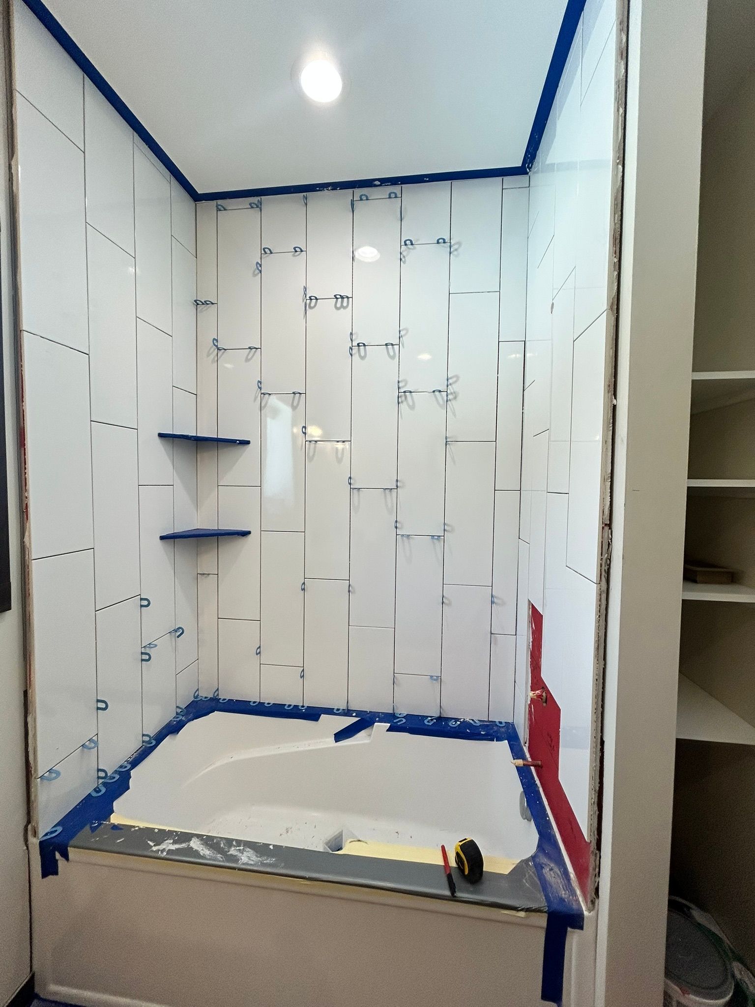A bathtub alcove under renovation with white tiled walls, blue painter's tape, and two installed corner shelves.