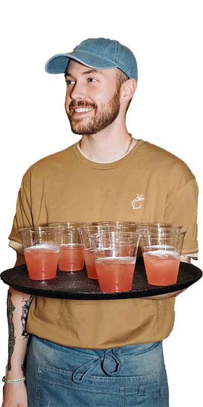 Man holding a tray with several red drinks, wearing a blue cap and tan shirt, smiling against a white background