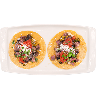 Two tacos on a white platter, topped with diced tomatoes, greens, and cheese