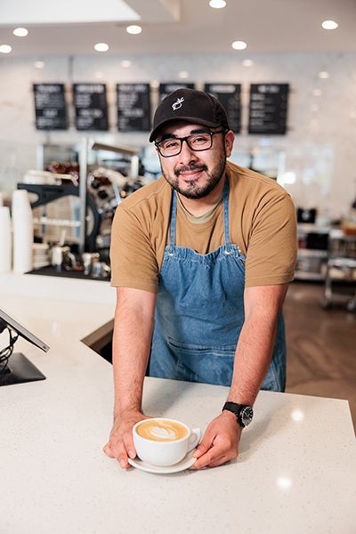 Barista in blue apron smiling behind a counter, holding a cup of coffee in a café.