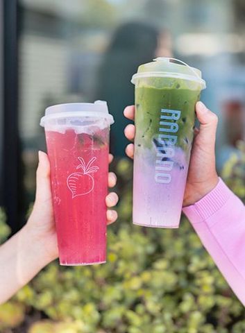 Two hands holding pink and green iced drinks outdoors at a cafe