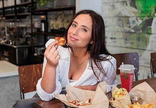 Woman smiling at a café table, holding a bite of food with drinks and takeout containers nearby