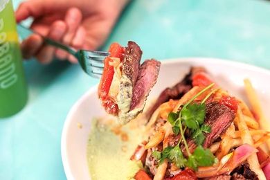 Fork lifting steak and pasta from a white bowl on a turquoise table