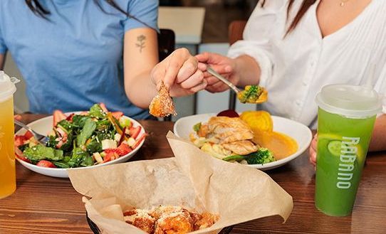 Two people sharing a meal with salad, pasta, fried food, and green drinks on a wooden table