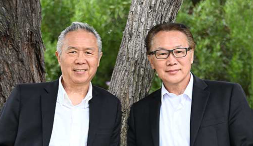 Two men in black jackets standing outdoors in front of tree trunks, smiling at the camera.