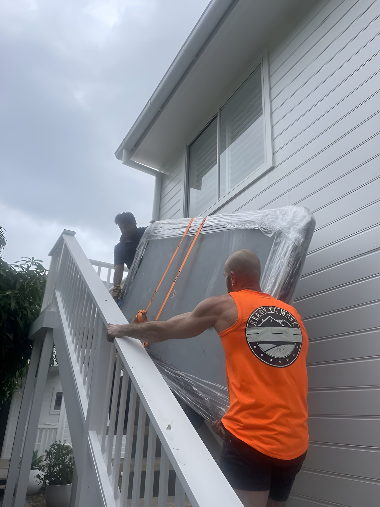 Two people carrying a large, wrapped item up outdoor stairs next to a white house— Ready To Move in Mullumbimby, NSW